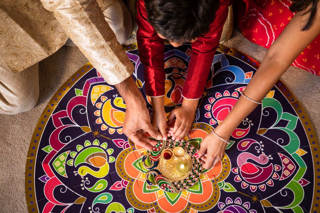 Hands placing a lit diya at the center of a colorful rangoli floor puzzle for Diwali