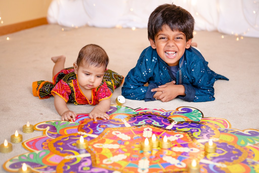 Baby and big brother in Indian clothes lying next to a rangoli floor puzzle with LED diyas for Diwali